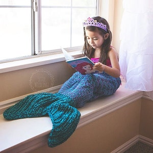 Op de afbeelding: Een kind met een paarse tiara en een blauw-groene zeemeerminstaartdeken leest een boek met een Ariel-illustratie terwijl het op een vensterbank zit. Het boek is open en het kind is geconcentreerd op de pagina's.