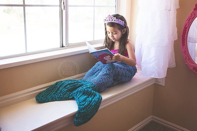 Op de afbeelding: Een kind met een paarse tiara en een blauw-groene zeemeerminstaartdeken leest een boek met een Ariel-illustratie terwijl het op een vensterbank zit. Het boek is open en het kind is geconcentreerd op de pagina's.