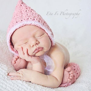 Puede incluir: Un gorro de bebé de ganchillo rosa con un borde de encaje blanco. El gorro lo lleva un bebé dormido con un calienta piernas de ganchillo rosa en una pierna. El bebé está tumbado en una manta blanca y esponjosa.