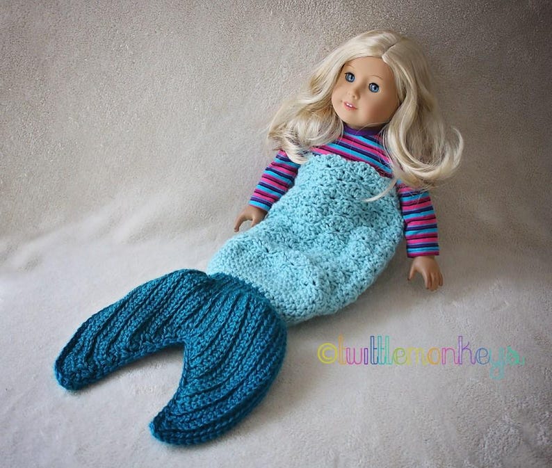 Op de afbeelding: Een pop gekleed in een handgemaakt gehaakt zeemeermin outfit. De pop heeft blond haar en blauwe ogen. De zeemeermin outfit is lichtblauw met een donkerblauwe staart. De bovenkant van de outfit heeft roze en blauwe strepen.