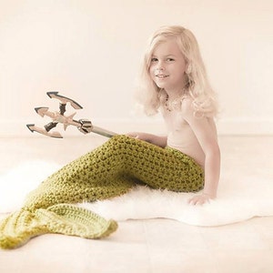 May include: A young person wearing a green crocheted mermaid tail sits on a white fluffy rug. They are holding a toy trident with a silver handle and black tines.