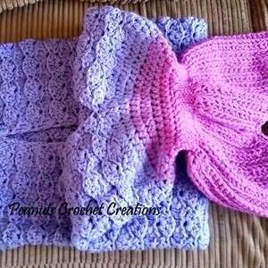 Op de afbeelding: Een handgemaakte gehaakte zeemeerminstaartdeken in tinten paars en lavendel. De deken heeft een gedetailleerd schubbenpatroon en een felroze staartvin. De tekst "Peanuts Crochet Creations" is zichtbaar.