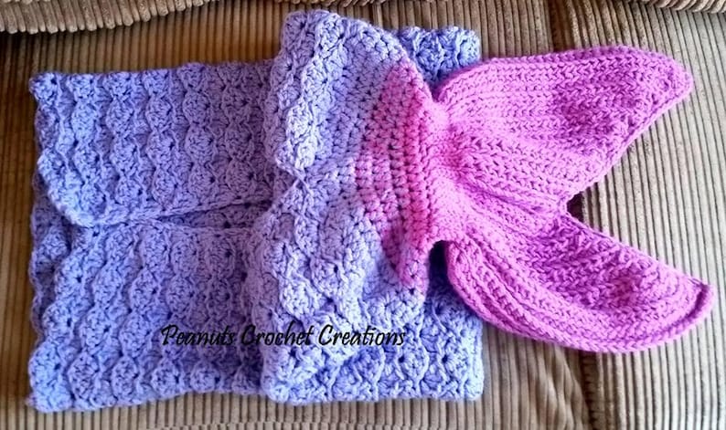 Op de afbeelding: Een handgemaakte gehaakte zeemeerminstaartdeken in tinten paars en lavendel. De deken heeft een gedetailleerd schubbenpatroon en een felroze staartvin. De tekst "Peanuts Crochet Creations" is zichtbaar.