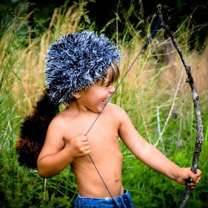 May include: A young person wearing a blue and white furry hat with a brown tail, holding a wooden bow and arrow. The person is standing in a grassy field.