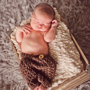 Puede incluir: Un bebé recién nacido durmiendo en una caja de madera con una manta blanca. El bebé lleva pantalones de punto marrones con un botón.