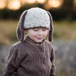 May include: A child wearing a handmade crochet hat with long, floppy ears. The hat is cream-colored with brown accents. The child is also wearing a brown zip-up sweater. The background is blurred with warm tones.
