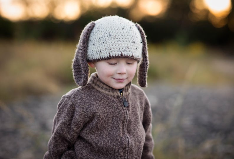 May include: A child wearing a handmade crochet hat with long, floppy ears. The hat is cream-colored with brown accents. The child is also wearing a brown zip-up sweater. The background is blurred with warm tones.