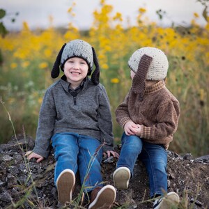 May include: Two children wearing gray and tan dog ear hats, gray and brown sweaters, and blue jeans. The children are sitting on a pile of rocks in a field of yellow flowers. The hats have long floppy ears.