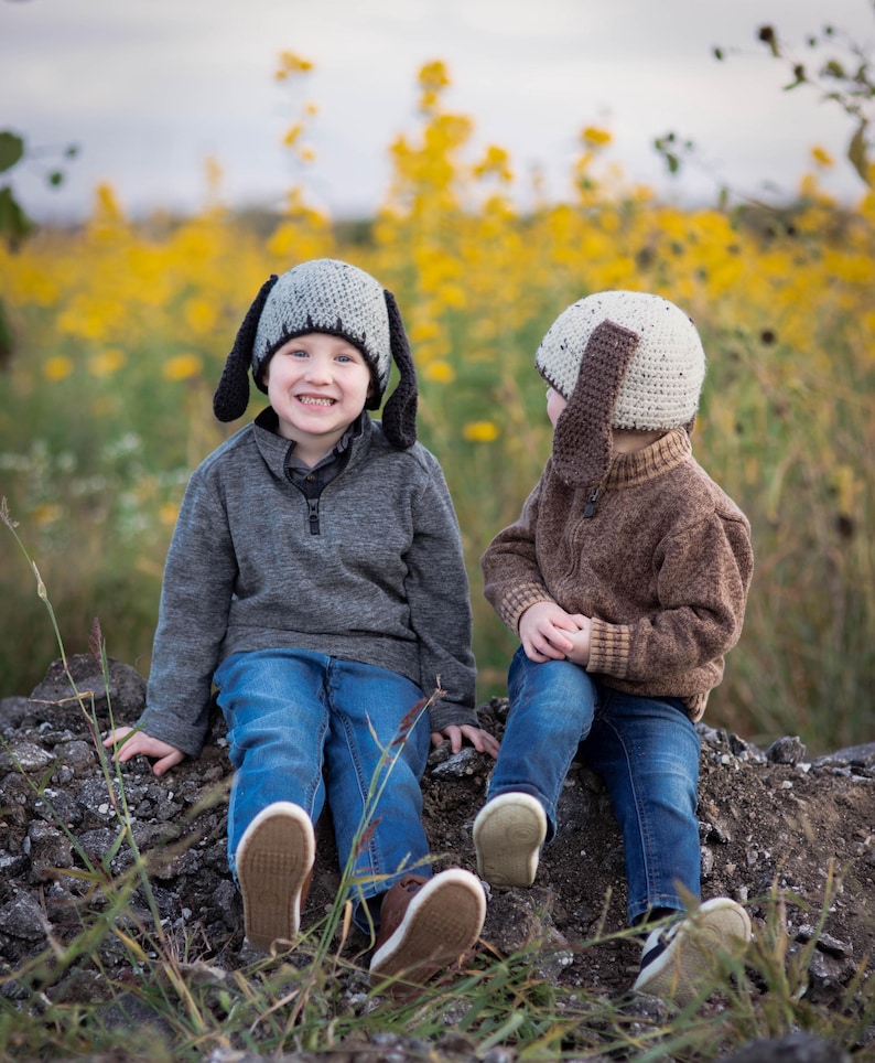 May include: Two children wearing gray and tan dog ear hats, gray and brown sweaters, and blue jeans. The children are sitting on a pile of rocks in a field of yellow flowers. The hats have long floppy ears.