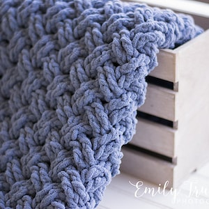 May include: A soft, blue knitted blanket draped over a wooden crate. The blanket has a textured, chunky knit pattern.