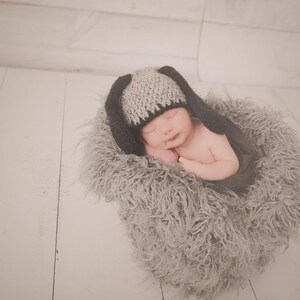 May include: A newborn baby wearing a gray and black bunny hat, nestled in a fluffy gray nest. The baby is sleeping peacefully, with arms gently folded. The background is a light-colored wooden surface.