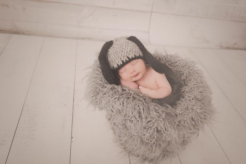May include: A newborn baby wearing a gray and black bunny hat, nestled in a fluffy gray nest. The baby is sleeping peacefully, with arms gently folded. The background is a light-colored wooden surface.