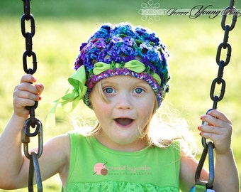 Puede incluir: Una niña pequeña lleva un gorro de ganchillo de colores con un lazo verde y está sentada en un columpio. Sonríe y mira a la cámara. El gorro está hecho con hilo de varios colores de morado, azul y verde. El texto "crochet by janelin crochet patterns" es visible en la parte delantera de su camiseta.