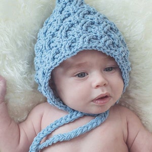 Peut inclure: Un bébé portant un bonnet en crochet bleu clair avec une cravate assortie. Le bonnet a un sommet pointu et une cravate tressée. Le bébé est allongé sur une surface blanche et moelleuse.