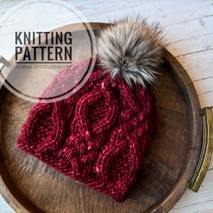 May include: A deep red chunky knit beanie featuring a cable knit pattern and a large, fluffy faux fur pom-pom on top. The image includes the text KNITTING PATTERN JENNIFERDOUGHERTY overlaid on the left side of the hat.