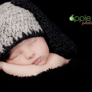 May include: A sleeping baby wearing a handmade crochet bunny hat with long ears. The hat is two-toned, with a light gray top and black trim. The baby is wrapped in a black blanket against a black background.