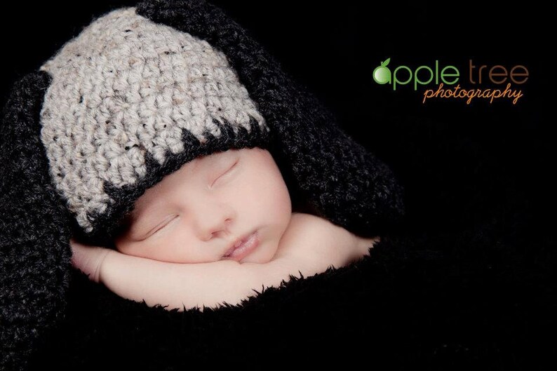 May include: A sleeping baby wearing a handmade crochet bunny hat with long ears. The hat is two-toned, with a light gray top and black trim. The baby is wrapped in a black blanket against a black background.