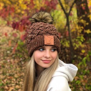 K&ouml;nnte beinhalten: Braune Strickm&uuml;tze mit einem Kunstfellbommel und einem Lederpatch mit der Umrandung des Bundesstaates Minnesota und dem Wort "Minnesota" darauf. Die M&uuml;tze wird von einer Person mit langen blonden Haaren getragen.