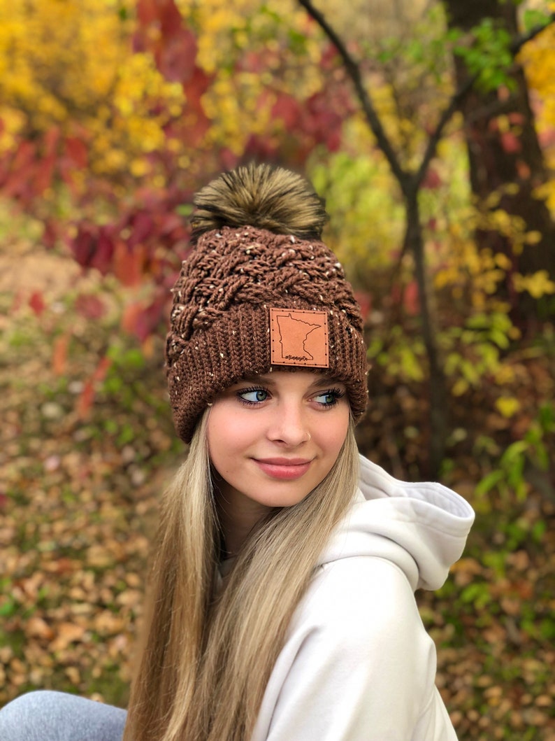 K&ouml;nnte beinhalten: Braune Strickm&uuml;tze mit einem Kunstfellbommel und einem Lederpatch mit der Umrandung des Bundesstaates Minnesota und dem Wort "Minnesota" darauf. Die M&uuml;tze wird von einer Person mit langen blonden Haaren getragen.