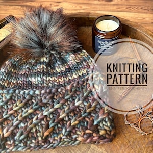 May include: Knitting pattern for a colorful beanie with a fluffy pom pom. The pattern is by Jennifer Dougherty.