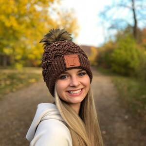 K&ouml;nnte beinhalten: Eine braune Strickm&uuml;tze mit einem Kunstfellbommel und einem Lederpatch mit der Umrissform des Bundesstaates Minnesota. Die M&uuml;tze wird von einer Person mit langen blonden Haaren getragen.
