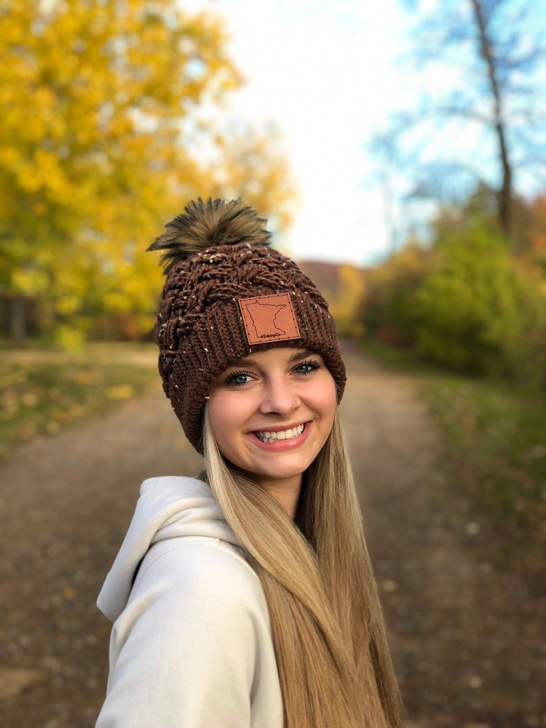 K&ouml;nnte beinhalten: Eine braune Strickm&uuml;tze mit einem Kunstfellbommel und einem Lederpatch mit der Umrissform des Bundesstaates Minnesota. Die M&uuml;tze wird von einer Person mit langen blonden Haaren getragen.