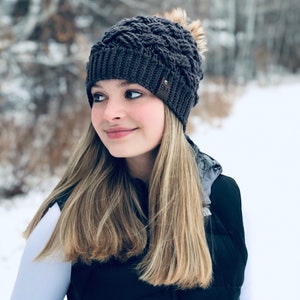 K&ouml;nnte beinhalten: Eine Frau tr&auml;gt eine graue Strickm&uuml;tze mit einem Bommel oben. Die M&uuml;tze hat einen braunen Lederfleck mit dem Wort "ORLJ" darauf. Sie tr&auml;gt eine schwarze Weste &uuml;ber einem wei&szlig;en Hemd.