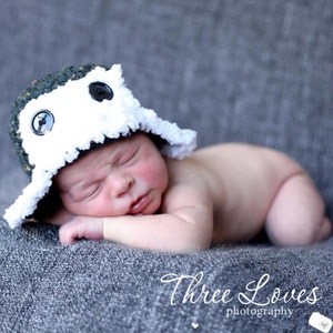 May include: A newborn baby wearing a handmade crochet hat with a white and dark green design, accented with black buttons. The baby is lying on a grey textured surface. The text "Three Loves photography" is visible.