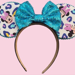 May include: A headband featuring Minnie Mouse ears with a teal sequined bow. The ears have a white background with a leopard print pattern in blue, purple, and yellow, and a Minnie Mouse graphic. The edges of the ears are trimmed with teal beads.