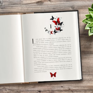 Puede incluir: Libro abierto con una página que presenta un diseño de mariposas. El diseño incluye mariposas negras y rojas, el número "1" y texto decorativo. El libro está sobre una superficie de madera.