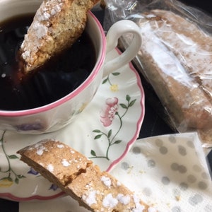May include: A cup of dark coffee with a biscotti dipped in it. The cup and saucer have a floral design with pink trim. Another biscotti rests on a polka-dot napkin, and a package of biscotti is in the background.