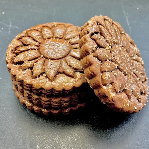 May include: Close-up of two chocolate cookies with a sunflower design. The cookies are stacked on top of each other and have a light brown color.