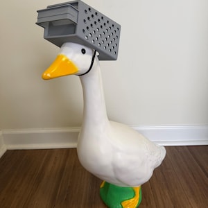 May include: A whimsical white goose statue with a yellow beak and feet, wearing a gray plastic crate on its head. The goose stands on a green base with yellow accents. The statue is a unique decorative item.