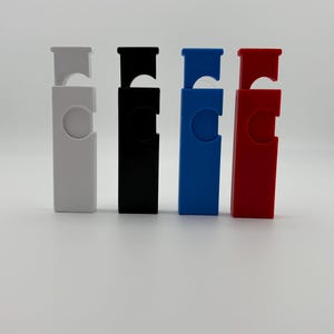 Puede incluir: Cuatro abridores de botellas de colores blanco, negro, azul y rojo. Cada abridor tiene un cuerpo rectangular con un recorte circular y una tapa con bisagras. Los abridores están dispuestos en fila sobre un fondo blanco.