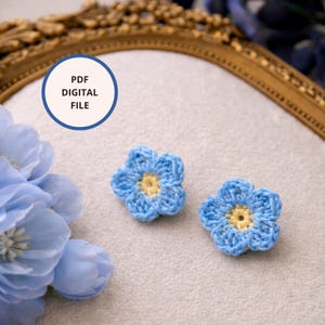 Puede incluir: Dos apliques de flores azules de ganchillo con centros amarillos. Las flores están sobre una superficie de color claro, con un marco decorativo dorado en el fondo. Un icono de archivo digital está en la esquina superior izquierda.