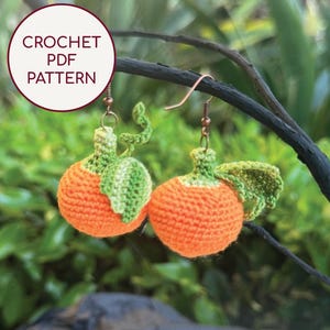 Op de afbeelding: Gehaakte oranje oorbellen met groene bladeren en koperkleurige oorhaken. De handgemaakte oorbellen hebben een getextureerd, driedimensionaal ontwerp. De afbeelding bevat de tekst "CROCHET PDF PATTERN" in een witte cirkel.