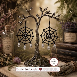 May include: A pair of black crocheted spider web earrings featuring a central white bead and three clear teardrop-shaped crystal dangles hanging from the bottom. The earrings are displayed on a metallic tree-shaped stand with a digital file label.