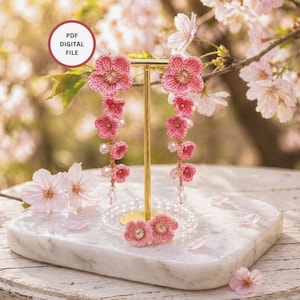May include: Pink crochet flower earrings with dangling flower accents and crystal beads. The earrings are displayed on a gold stand, with a matching flower brooch at the base. The background features cherry blossoms and the text "PDF DIGITAL FILE".