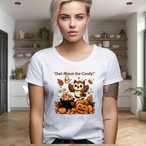 Może przedstawiać: Biała koszulka z motywem Halloween. Projekt przedstawia kreskówkową sowę, dynie, cukierki i napis "Owl About the Candy!". Sowa jest brązowo-biała, z pomarańczowymi akcentami. Dynie są pomarańczowe, a cukierki są żółte, pomarańczowe i białe.