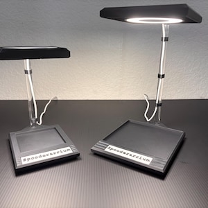 May include: Two black desk lamps with a square base and a rectangular light. The lamps have a clear, cylindrical stem and a white cord. The text "spooderrarium" is printed on the base of each lamp.