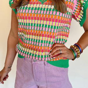 Könnte beinhalten: Ein kurzärmeliges Strickoberteil mit einem lebendigen geometrischen Muster in Grün, Pink, Orange und Gelb, mit V-Ausschnitt und grünem Rippstricksaum, kombiniert mit hellvioletten Jeansshorts, die zwei große aufgesetzte Vordertaschen haben.