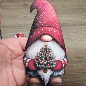 Può includere: Statuetta di gnomo natalizio con cappello rosso a pois bianchi e lunga barba bianca. Lo gnomo tiene in mano un albero di Natale decorato con luci e palline. Lo gnomo indossa guanti rossi e un cappotto blu.