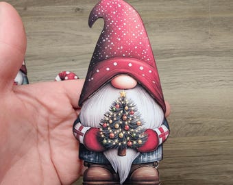 May include: A Christmas gnome figurine with a red hat dotted with white spots and a long white beard. The gnome is holding a decorated Christmas tree with lights and ornaments. The gnome is wearing red mittens and a blue coat.