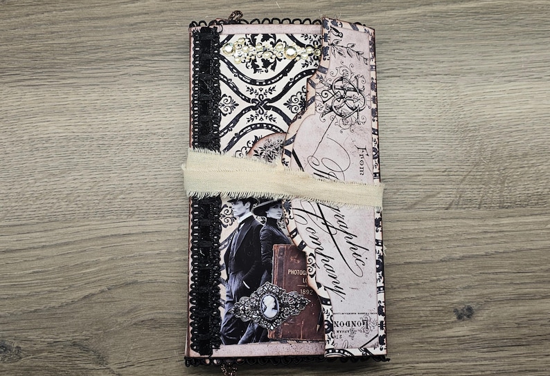 May include: A vintage-style journal with a black and white damask pattern, embellished with black lace trim and a cream-colored ribbon. The cover features a sepia-toned image of a couple and decorative elements.
