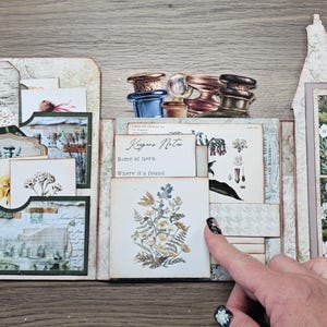 May include: A paper craft journal featuring botanical illustrations, vintage-style tags, and pockets. The center card displays the text Keepers Notes, Name of herb, and Where it's found, accompanied by a floral print. Includes apothecary bottle imagery.