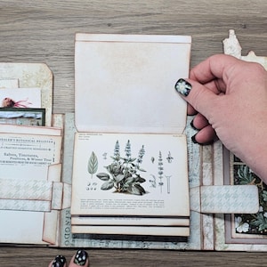 May include: A handcrafted paper journal featuring a botanical illustration of a plant with green leaves and stems. The page includes printed text titled The Healer's Botanical Register, Salves, Tinctures, and Winter Teas, with additional paper inserts.
