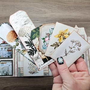 May include: A collection of paper ephemera including rectangular tags and cards featuring botanical illustrations of yellow flowers, white blossoms, and green foliage, alongside a multi-pocketed folder with apothecary bottle graphics and text.