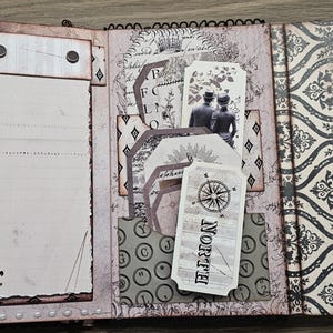 May include: Open scrapbook with vintage-style pages. Features include lined paper, decorative tags with a compass rose and couple illustration, and patterned paper with a damask design. The color palette is primarily neutral with accents of black.