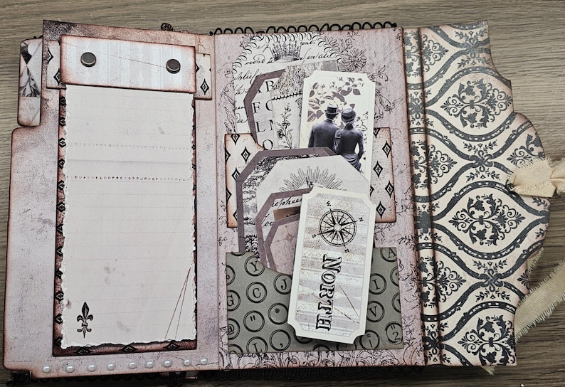 May include: Open scrapbook with vintage-style pages. Features include lined paper, decorative tags with a compass rose and couple illustration, and patterned paper with a damask design. The color palette is primarily neutral with accents of black.