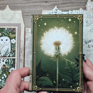May include: A rectangular book with a dark green cover featuring a white dandelion seed head, fern leaves, and gold corner accents. The background displays a printed scene of a white owl and a glass bottle with botanical illustrations.